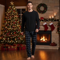 A man stands barefoot in a cozy holiday living room wearing a black long-sleeve crewneck shirt and dark blue plaid pajama pants. A decorated Christmas tree with warm lights, a fireplace with stockings, and wrapped gifts are visible in the background.