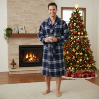 A man wearing a blue and white plaid bathrobe stands barefoot in a living room in front of a fireplace and a decorated Christmas tree.