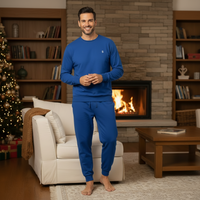 A man wearing a matching royal blue long-sleeve crewneck shirt and jogger pants stands barefoot in a cozy living room in front of a fireplace. The room features a white armchair, a wooden coffee table, bookshelves, and a Christmas tree on the left.