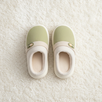A pair of slip-on indoor slippers with a light sage green ribbed upper, cream ribbed collar, and white soles, laid flat on a white textured rug.
