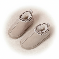 A pair of beige slip-on baby booties with white faux-fur lining and a decorative zigzag stitched trim around the collar, displayed on a soft white textured surface.