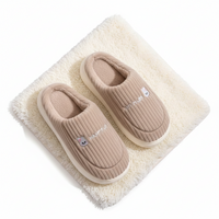 A pair of beige ribbed slip-on house slippers with a small bear face graphic and the word 'NININU' on the side, displayed on a fluffy cream rug against a white background.