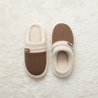 A pair of slip-on indoor slippers shown from above on a white faux-fur surface. Each slipper has a brown ribbed knit lower section, a cream ribbed upper band, a cream padded collar and footbed, and a small oval brand patch on the side.