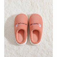 A pair of pink ribbed slip-on house slippers with white soles, placed on a white textured rug. Each slipper has a small embroidered cartoon face and the text 'HAPPY LIFE' in black.