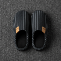 A pair of black slip-on slippers with thin white vertical pinstripes and tan brand patches, photographed from above on a dark gray textured carpet.