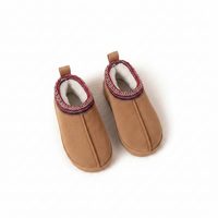 A pair of tan slip-on baby booties with a soft white fleece lining and a red and navy patterned trim around the collar, shown on a white background.