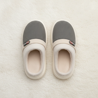 A pair of slip-on indoor slippers shown from above on a soft white textured surface. The slippers have gray ribbed uppers, cream knit trim, cream footbeds, and cream soles.
