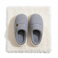A pair of light gray ribbed slip-on house slippers with white soles, displayed on a white shag rug. The left slipper features a small brown bear face logo on the side.