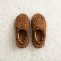 A pair of cozy slip-on slippers in a warm camel brown sherpa fabric, laid flat on a white faux fur rug.