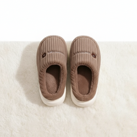 A pair of cozy brown corduroy slip-on slippers with white soles, photographed from above on a white textured rug.