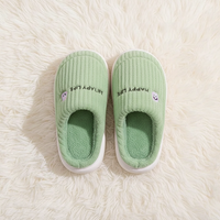 A pair of light green ribbed slip-on slippers with white soles, placed on a white faux fur rug. The slippers feature the text 'HAPPY LIFE' in black and a small embroidered smiley face on the side.