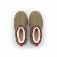 A pair of slip-on winter slippers shown from a top-down view on a white background. The slippers have a taupe-brown synthetic exterior, a red and beige patterned trim around the collar, a cream-colored fleece lining, and a small red pull tab.