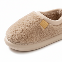 A close-up of a beige sherpa fleece slip-on house slipper with a tan brand patch on the side, set against a clean white background.