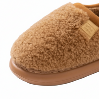 A close-up of a tan sherpa fleece slipper with a tan rubber sole and a small tan leather brand patch that reads 'WITNESS HOME'.