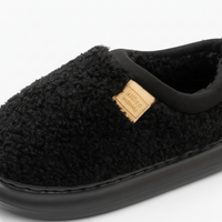 A close-up of a black fluffy fleece slipper with a tan leather brand patch that reads 'NILES HOMME' on a white background.
