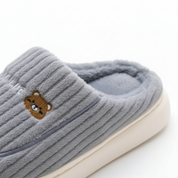 A close-up of a gray ribbed fleece slipper with a small embroidered brown bear face on the side and a cream-colored sole, shown on a white background.
