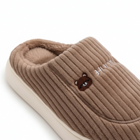 A close-up of a ribbed brown slip-on slipper with a plush lining, a small embroidered bear face on the side, and white lettering, shown on a white background.