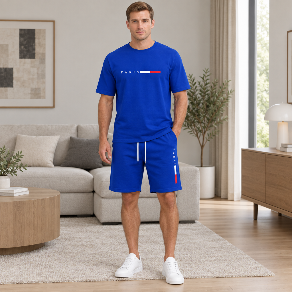 A man stands in a modern living room wearing a royal blue matching set consisting of a short-sleeve T-shirt and shorts. The shirt features 'PARIS' in white above a red, white, and blue flag motif, and the shorts have white drawstrings with a vertical red, white, and blue stripe on the side.