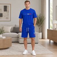 A man stands in a modern living room wearing a royal blue matching set consisting of a short-sleeve T-shirt and shorts. The shirt features 'PARIS' in white above a red, white, and blue flag motif, and the shorts have white drawstrings with a vertical red, white, and blue stripe on the side.