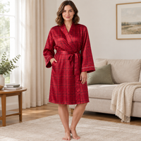 A woman stands barefoot in a bright living room wearing a red plaid bathrobe with a tie waist and long sleeves. The robe has a subtle sheen and is shown in a warm, well-lit interior setting with a sofa, side table, and curtains in the background.