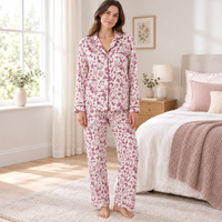 A woman stands in a bright bedroom wearing a long-sleeve pajama set with a pink and burgundy floral print on a white background. The set includes a button-front top and matching pants.