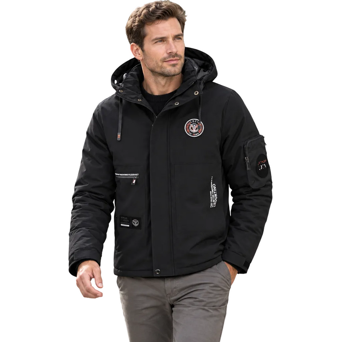 A man wearing a black hooded winter jacket with multiple pockets and a circular patch on the chest, walking on a city sidewalk.