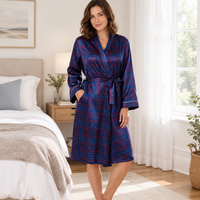 A woman wearing a long-sleeve plaid bathrobe in a bedroom setting. The robe features a blue, purple, and red tartan pattern with white piping on the collar, cuffs, and waist tie belt.