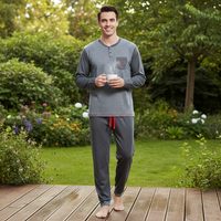 A man stands barefoot on a wooden deck in a lush garden, wearing a gray long-sleeve striped henley pajama top with a red drawstring and matching gray pants.