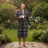 A man wearing a black and white plaid bathrobe stands barefoot on a wooden deck in a lush garden, holding a white mug.