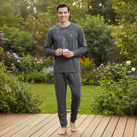A man wearing a dark gray long-sleeve pajama shirt and matching jogger pants stands barefoot on a wooden deck in a lush garden, holding a white mug.
