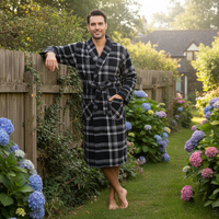 A man wearing a black and white plaid bathrobe stands barefoot in a lush garden pathway, leaning against a wooden fence with hydrangeas in the background.