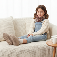 A young girl sits on a cream-colored textured couch, wearing a cream cable-knit sweater, light blue denim overalls, a brown and burgundy patterned knit scarf, and taupe slip-on shoes. A small wooden side table with white mugs is visible on the right.
