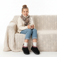 A young girl sits on a cream cable-knit sofa, wearing a cream knit sweater, a brown and beige patterned scarf, light blue jeans, white socks, and black slip-on boots. She is holding a white mug against a clean white background.
