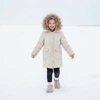 A young child smiling while walking in the snow, wearing a light beige quilted winter parka with a brown faux-fur trimmed hood, dark pants, and knit boots.