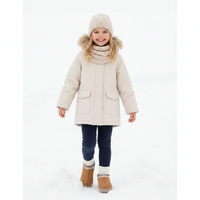 A young girl smiling in a snowy outdoor setting, wearing a light beige winter parka with a faux-fur trimmed hood, a matching knit beanie, a knit neck warmer, dark blue leggings, and tan boots.