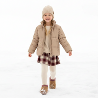 A young girl smiling in a snowy outdoor setting, wearing a beige puffer jacket, cream knit sweater, a red and brown plaid skirt, cream tights, and tan boots.