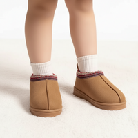 A pair of tan slip-on slippers with a multicolor patterned trim, shown on a doll's legs wearing white socks against a white background.