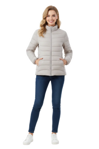 A woman modeling a light beige puffer jacket with a high collar and front zipper, paired with dark blue skinny jeans and white sneakers, against a plain white studio background.