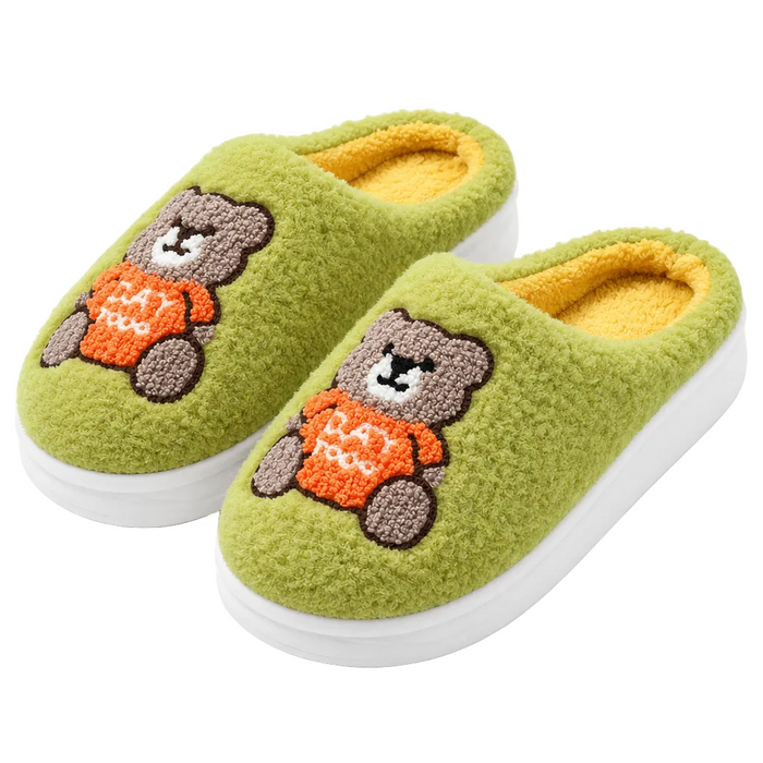 A pair of plush slip-on house slippers in light olive green, featuring a teddy bear motif on the upper. The bear is brown with a gray snout and wears an orange sweater with the word 'LOVE' in white.