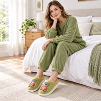 A woman sits on a bed wearing a matching sage green fleece pajama set with cuffed pants and slip-on slippers featuring a teddy bear graphic. The bedroom has white bedding, a wooden nightstand, and a potted plant.