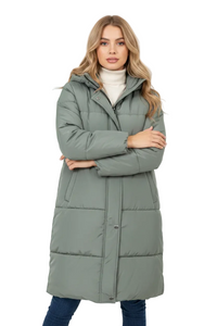 A woman modeling a long, sage green puffer coat over a white turtleneck sweater and blue jeans, standing against a plain white studio background.