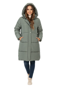 A woman modeling a long, hooded puffer coat in a muted sage green color, paired with dark blue jeans and beige sneakers, against a plain white studio background.