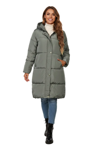 A woman modeling a mid-length, hooded puffer coat in a muted sage green color. She is wearing a white turtleneck, blue jeans, and black ankle boots, standing against a solid white background.