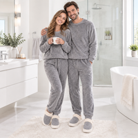 A smiling couple wearing matching gray textured fleece pajama sets with drawstring sweatshirts and pants, standing in a bright modern bathroom. The woman holds a white mug, and both wear gray slip-on slippers with white trim.