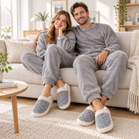 A couple sitting together on a light beige couch in a bright living room, wearing matching gray fleece lounge sets and quilted slip-on slippers with white trim.
