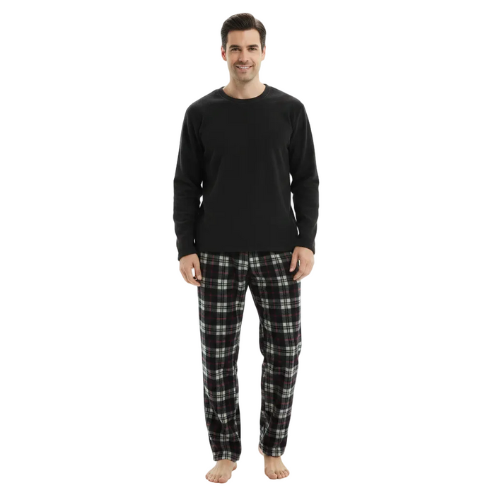 A man standing against a plain white background wearing a black long-sleeve crewneck shirt and black, white, and gray plaid pajama pants.