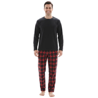 A man standing against a plain white background wearing a black long-sleeve crewneck shirt and red and black plaid pajama pants.