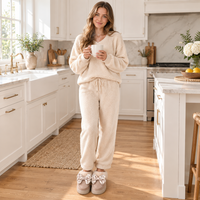 A woman stands in a bright white kitchen wearing a cozy cream fleece pajama set with matching slippers and holding a white mug.