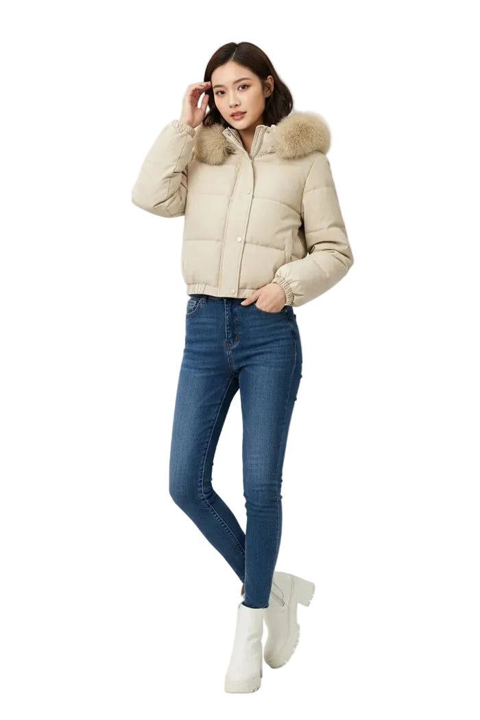 A woman modeling a beige puffer jacket with a fluffy faux-fur trimmed hood, paired with dark blue skinny jeans and white ankle boots, against a plain white studio background.