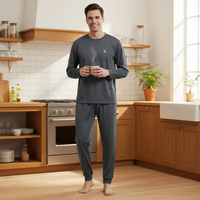 A man stands barefoot in a bright kitchen wearing a dark gray long-sleeve pajama set with matching pants. The pajama top has a small white embroidered logo on the left chest, and the set has a subtle steam graphic on the chest.